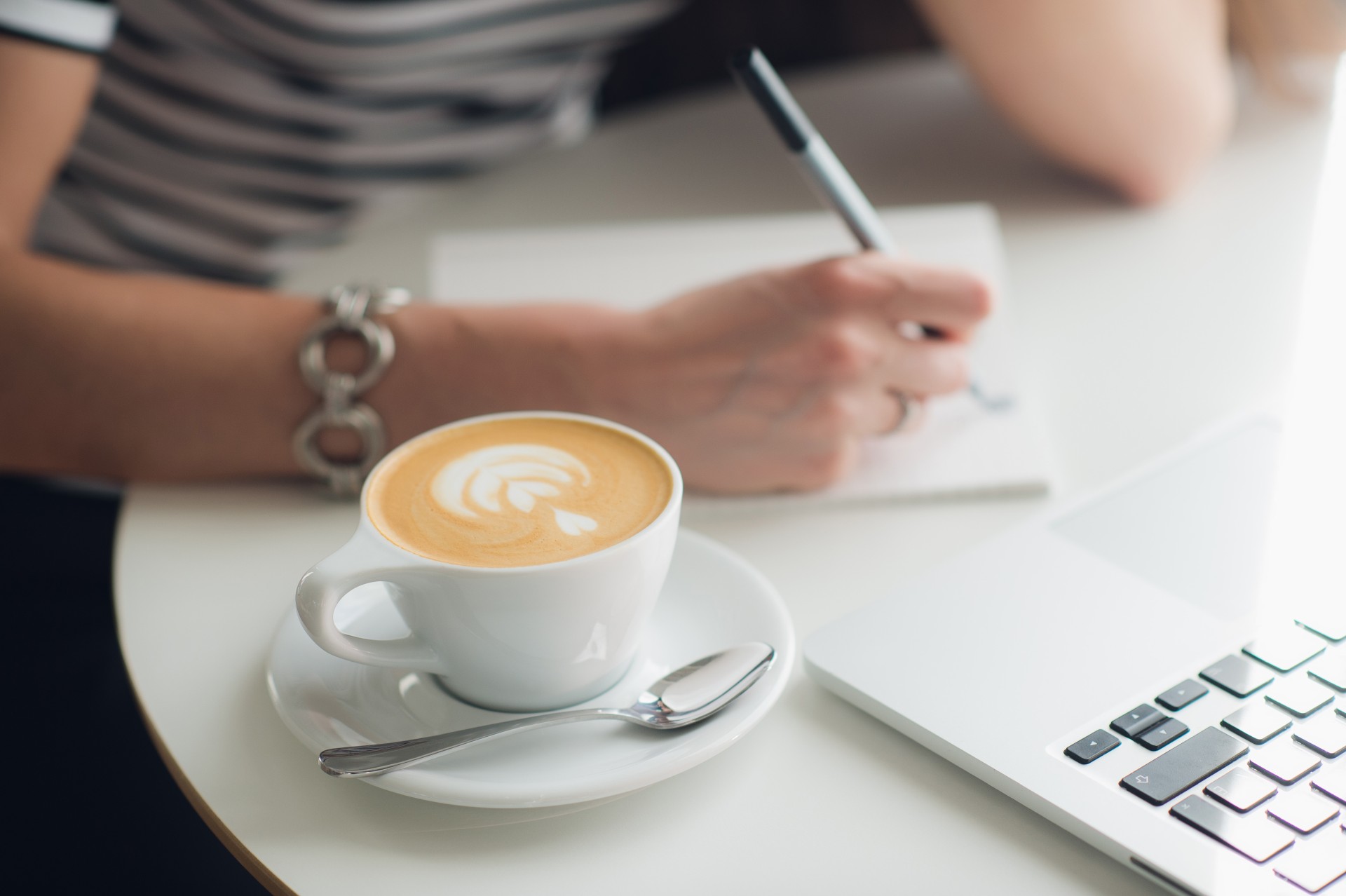 Close up picture of woman’s hands and a cup of cappuccino. Lady is writing in her notebook with a laptop nearby.